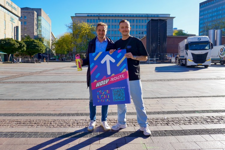 Carsten Hinzer, Brand Activation Lead bei HOLY und EMG-Gesch&auml;ftsf&uuml;hrer Richard R&ouml;hrhoff pr&auml;sentieren auf dem Kennedyplatz die neue Routenbeschilderung des Festivals.