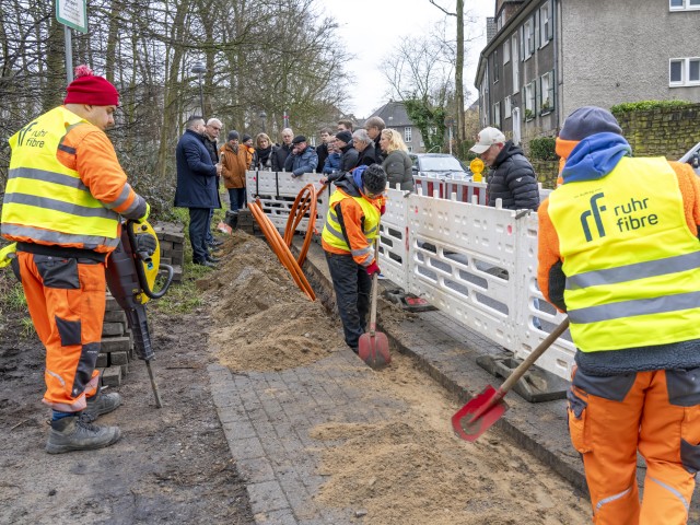 Foto: Bauarbeiten am Straßenrand, Bauarbeiter verlegen Kabel, unterstützende und interessierte Personen stehen an der Absperrung