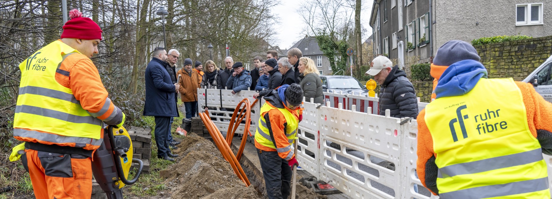 Foto: Bauarbeiten am Straßenrand, Bauarbeiter verlegen Kabel, unterstützende und interessierte Personen stehen an der Absperrung