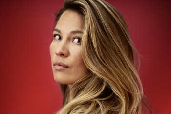 Eine Frau mit langen, hellbraunen Haaren blickt über die Schulter in die Kamera. Sie trägt ein schwarzes Oberteil und steht vor einem kräftig roten Hintergrund. Ihr Ausdruck ist ruhig und konzentriert, das Licht betont die Struktur ihres Haares und die warmen Hauttöne. Das Porträt wirkt klar, modern und aufmerksam komponiert.