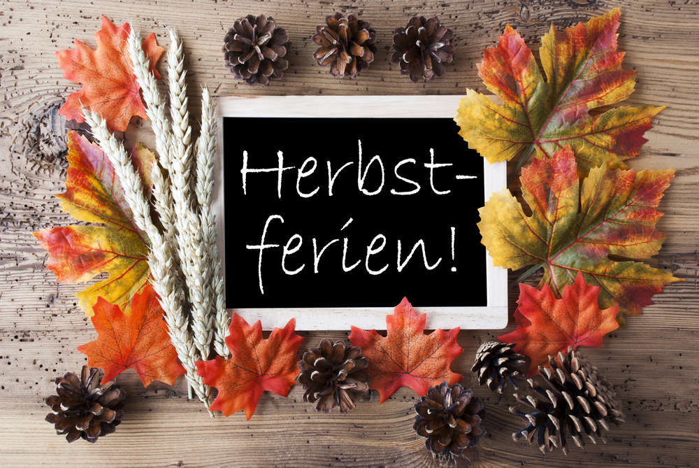 Auf einem Holztisch liegt eine kleine Tafel mit der weißen Aufschrift „Herbstferien!“. Sie ist von bunten, herbstlich verfärbten Blättern, Tannenzapfen und Ähren umgeben. Die Farben reichen von warmem Rot und Orange bis zu sattem Gelb und Braun. Das Arrangement vermittelt eine gemütliche, naturverbundene Herbststimmung.