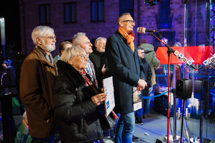Sprecher mit Gruppe am WDR-Mikrofon bei Weihnachtsaktion vor historischer Kulisse in blau-violettem B&uuml;hnenlicht
