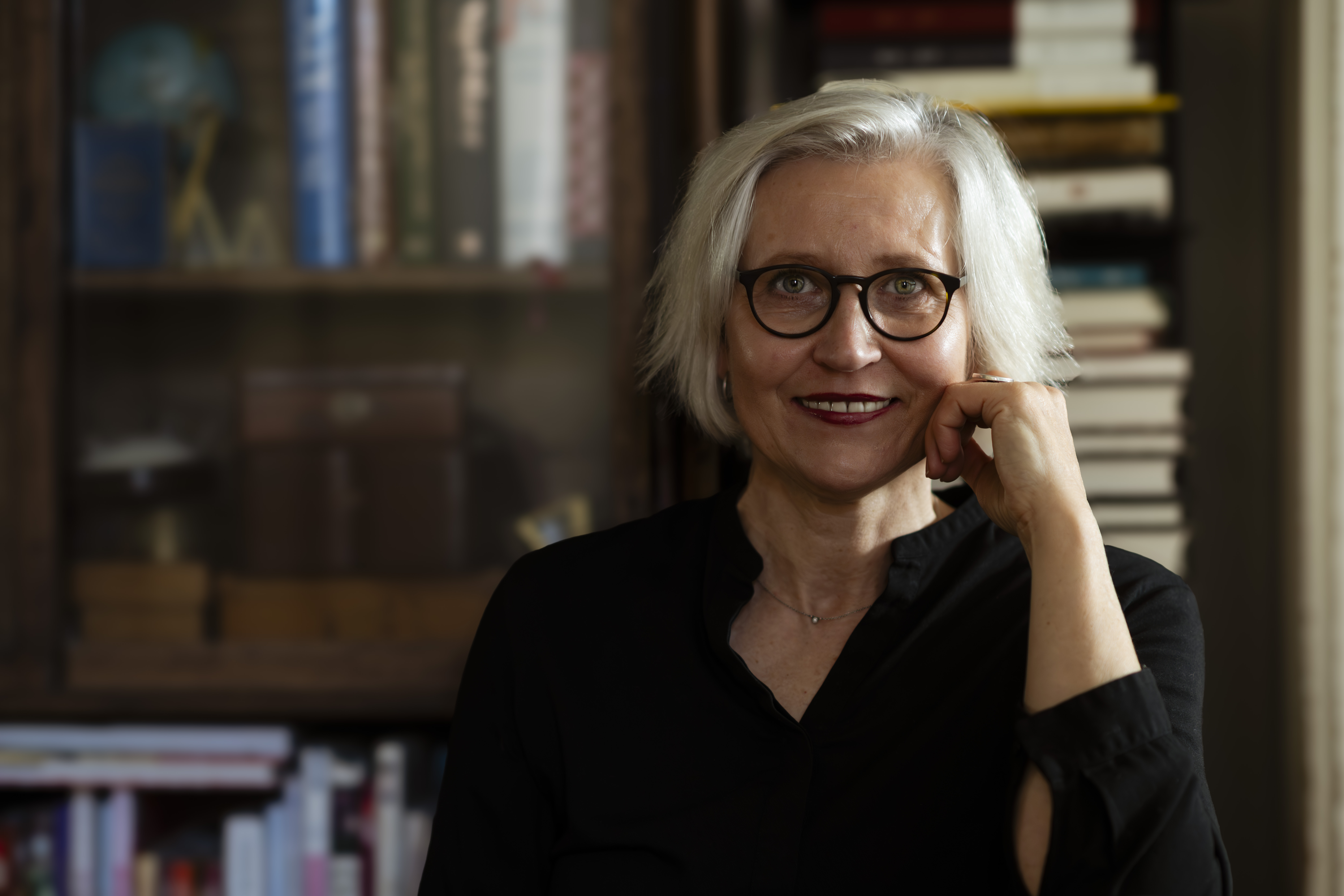 Portr&auml;t einer Frau vor einem B&uuml;cherregal. Sie blickt freundlich frontal in die Kamera. Ihr kurzes, helles Haar f&auml;llt leicht zur Seite. Sie tr&auml;gt eine schwarze Bluse und eine runde dunkle Brille. Eine Hand ist an die Wange gelegt. Der Hintergrund ist unscharf und zeigt B&uuml;cher sowie warme dunkle Holzt&ouml;ne.