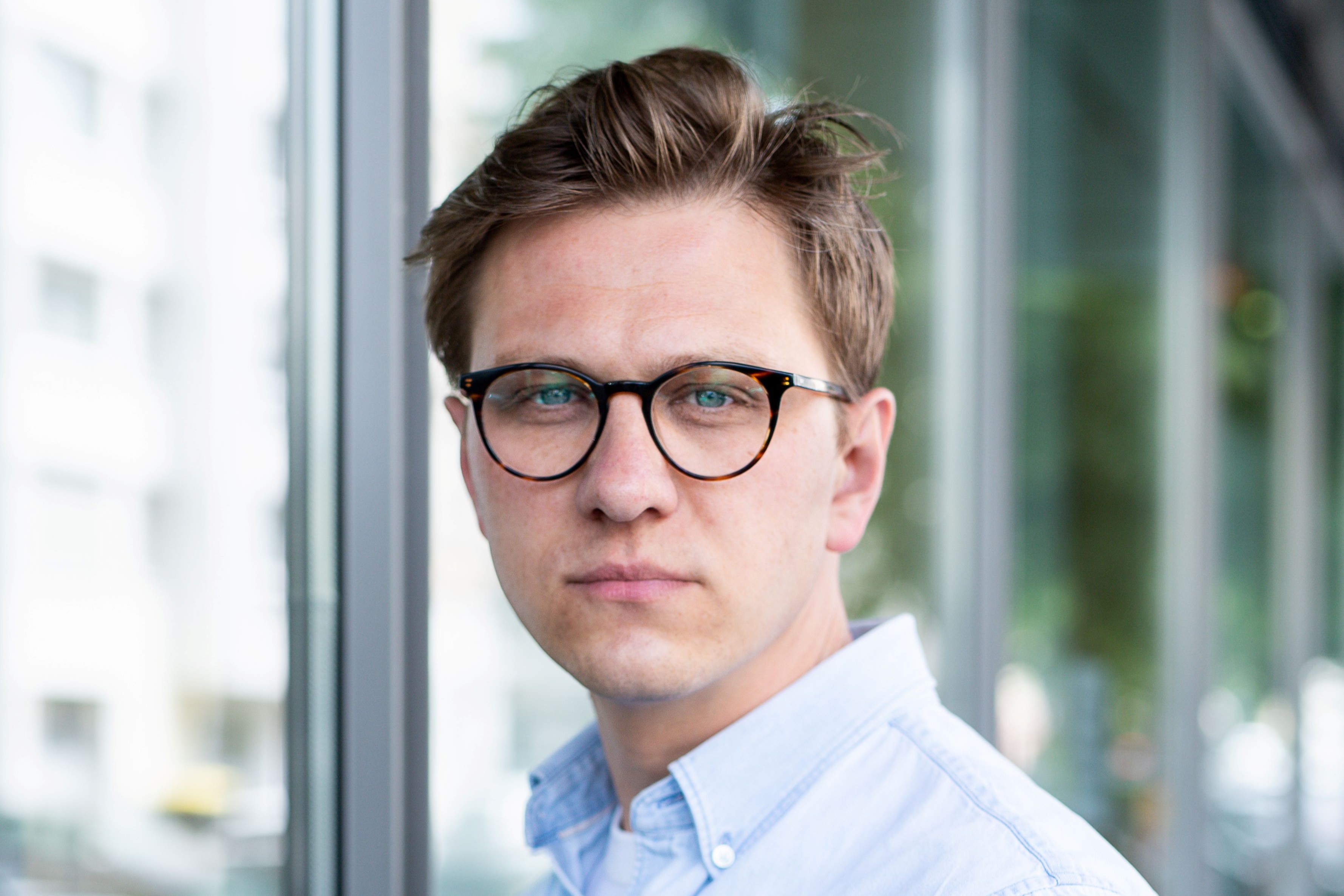 Ein junger Mann im blauen Hemd blickt ernst in die Kamera. Er tr&auml;gt ein blaues Hemd und hat braunes Haar.