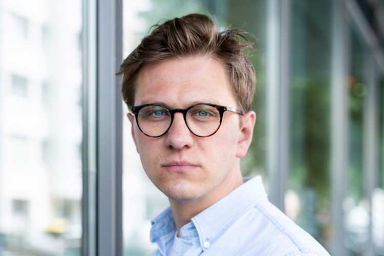 Ein junger Mann im blauen Hemd blickt ernst in die Kamera. Er tr&auml;gt ein blaues Hemd und hat braunes Haar.