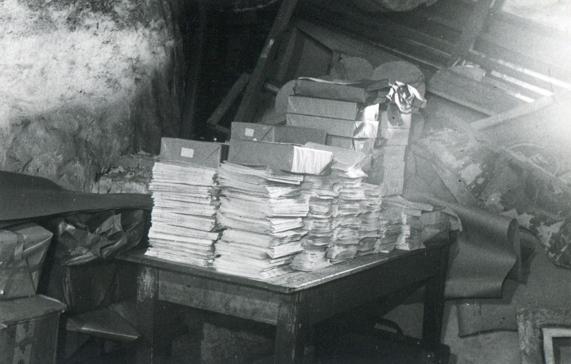 Zu sehen ist ein schwarz-wei&szlig;es Bild. In der Mitte eines chaotischen Abstellraums steht ein massiver Holztisch. Darauf stapeln sich Massen an Papieren und Kartons. Es handelt sich um konfiszierte illegale Druckschriften.