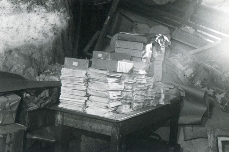 Zu sehen ist ein schwarz-wei&szlig;es Bild. In der Mitte eines chaotischen Abstellraums steht ein massiver Holztisch. Darauf stapeln sich Massen an Papieren und Kartons. Es handelt sich um konfiszierte illegale Druckschriften.