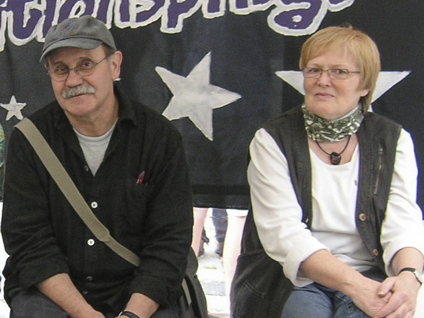 Zwei Erwachsene schauen in die Kamera: Der Mann sitzt links und tr&auml;gt eine runde Brille, eine graue Cap und einen grauen Schnurrbart. Die Frau hat kurze blonde Haare und eine viereckige Brille.