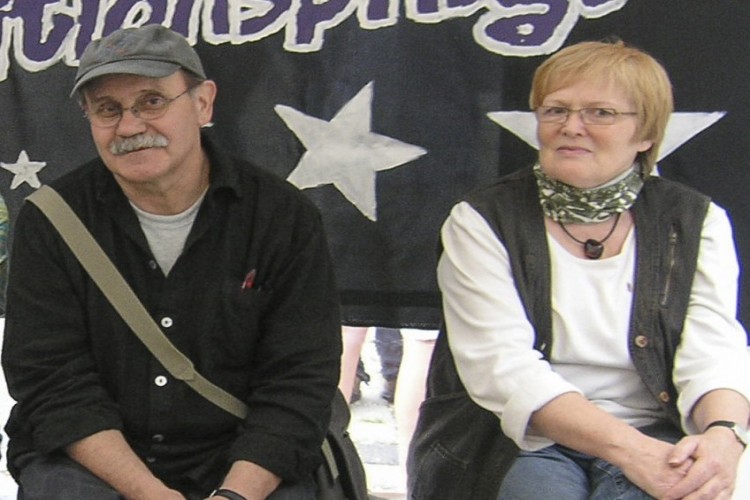 Zwei Erwachsene schauen in die Kamera: Der Mann sitzt links und tr&auml;gt eine runde Brille, eine graue Cap und einen grauen Schnurrbart. Die Frau hat kurze blonde Haare und eine viereckige Brille.