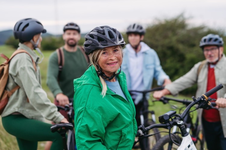 Eine Gruppe Menschen macht w&auml;hrend einer Fahrradtour eine Pause. Alle tragen Helme. Manche reden miteinander. Sie stehen bei ihren Fahrr&auml;dern. Eine Frau in neongr&uuml;ner Regenjacke steht im Vordergrund und schaut in die Kamera.
