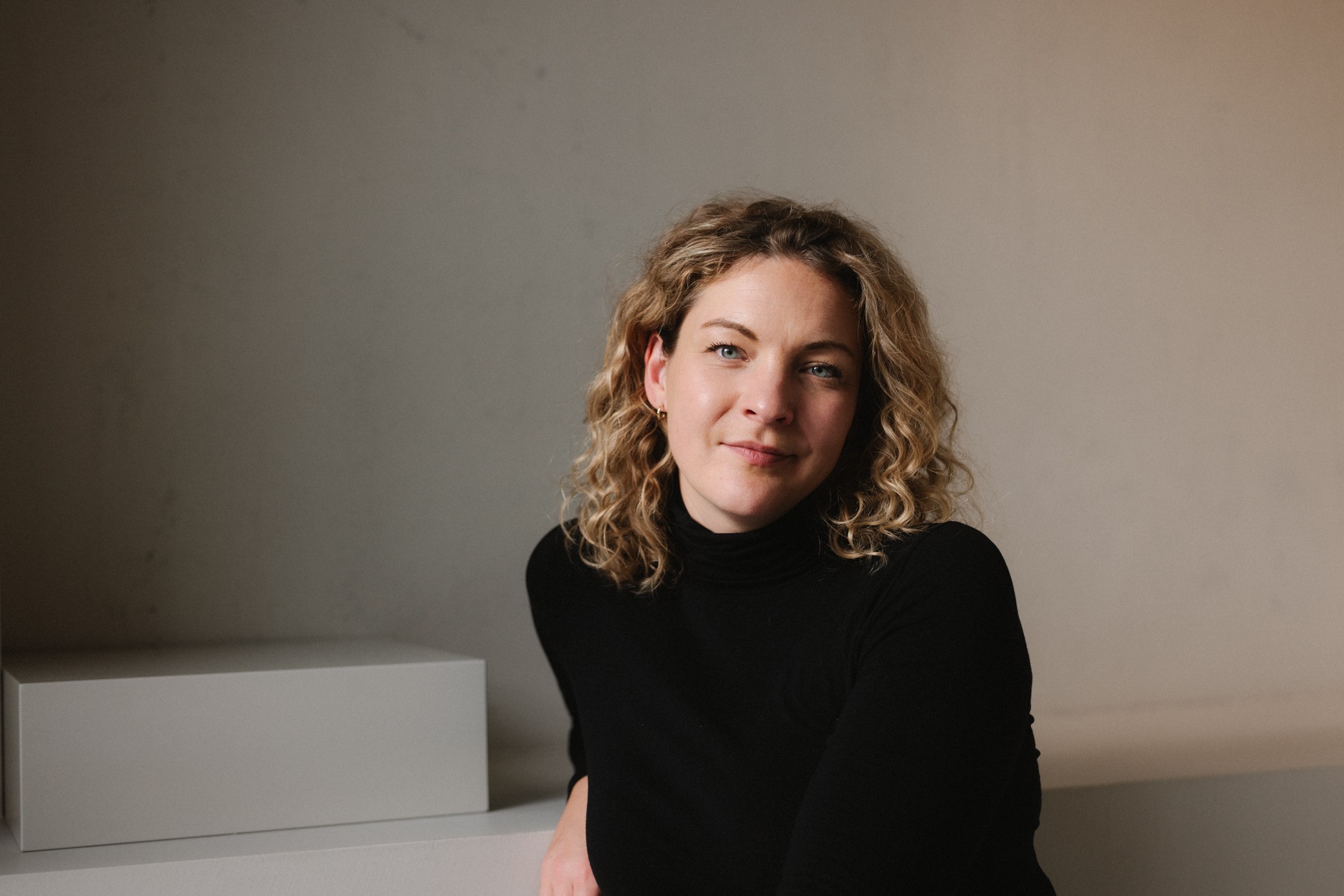 Eine junge Frau mit blonden Haaren und schwarzem Rollkragenpullover schaut in die Kamera.