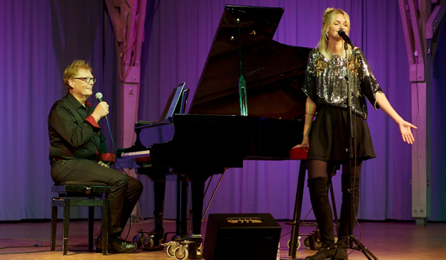 Ein Mann, Heinz Peter Reidmacher, sitzt an einem schwarzen Fl&uuml;gel und h&auml;lt ein Mikrofon in der Hand. Er ist schick gekleidet, mit schwarzer Hose und Hemd. Neben ihm steht eine Frau, Nora Mosch, mit langem blonden Haar und einem schicken Glitzeroberteil, Rock und Stiefeln. Sie singt mit geschlossenen Augen in ein Standmikrofon. 