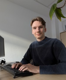 Studierender sitzt an seinem Laptop und guckt in die Kamera