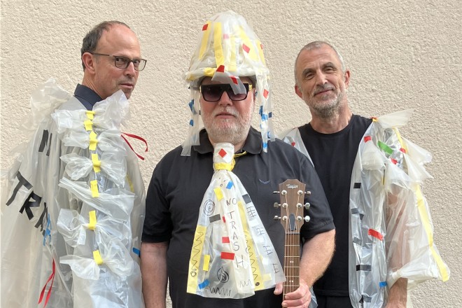 drei Männer in bunten Kostümen, der Mann in der Mitte hält eine Gitarre