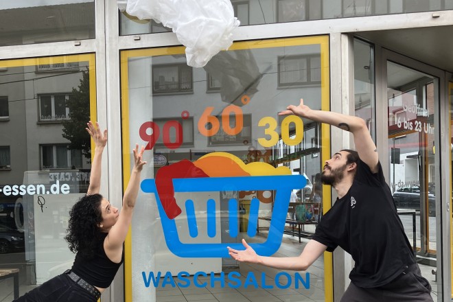 eine Frau und ein Mann in dunkler Tanzkleidung werfen vor einem Waschsalon eine Plastiktüte in die Luft
