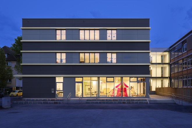 Das Foto zeigt die Fassadenansicht des Erweiterungsneubaus der Grundschule Sternschule an der Brigittastraße in Essen aus nordöstlicher Richtung am Abend. Das dreigeschossige Gebäude hat eine schwarze Fassade, die im zweiten und dritten Obergeschoss durch beigefarbig abgesetzte Fensterbänder unterbrochen wird. Die Innenbeleuchtung ist in den Räumen eingeschaltet. Im Erdgeschoss befindet sich der beleuchtete Mensabereich der Schule. Rechts im Bild ist das Bestandsgebäude der Schule zu sehen, dass durch einen dreigeschossigen Gebäuderiegel mit dem Neubau verbunden ist.