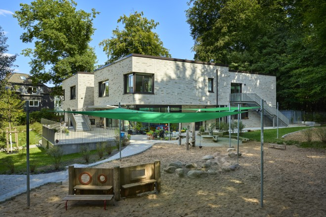 Das Foto zeigt den Neubau der zweigeschossigen Kindertagesstätte am Erikapfad in Essen-Stadtwald von Süd-West. Das Gebäude hat ein Flachdach und eine Fassade aus hellen Klinkersteinen. Links und rechts führen je eine Stahltreppe vom Obergeschoss des Gebäudes in den Außenbereich. Vor dem Neubau ist ein großzügiger Sandspielplatz mit Spielgeräten und zwei grünen Sonnenschutzschutzsegeln zu sehen. Markante Details des Gebäudes sind die über die Gebäudeecken verglasten Fenster.