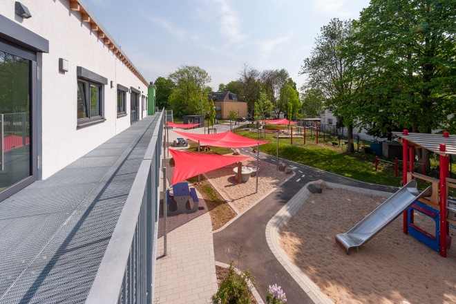 Foto: Das Foto zeigt den Blick vom Balkon aus südwestlicher Richtung auf die Außenanlagen. Hier ist ein Spiel- und Klettergerät mit Rutsche, ein Wassermatschbereich und Schaukeln sowie im Hintergrund der Sandspielbereich für unter dreijährige Kinder zu sehen.