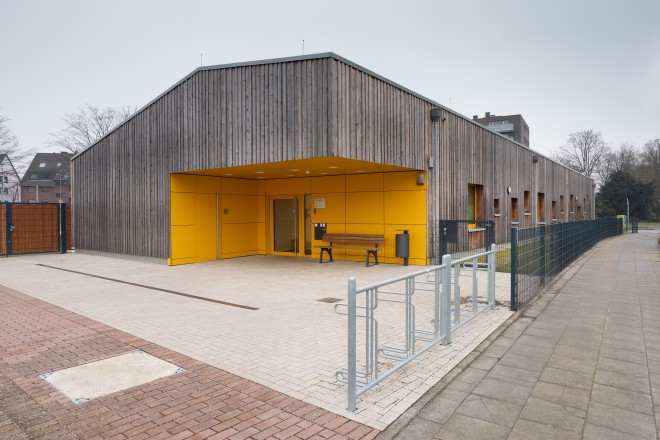Das Foto zeigt eine Gebäudeecke des eingeschossigen Neubaus der Kindertagesstätte an der Fulerumer Straße 100 in Essen-Haarzopf aus Südöstlicher Richtung. Die Fassade zeigt sich in einer Holzverkleidung, die durch verschieden große und farbige Fenster sowie einen auffällig in Gelb gehaltenen Haupteingangsbereich unterbrochen wird.