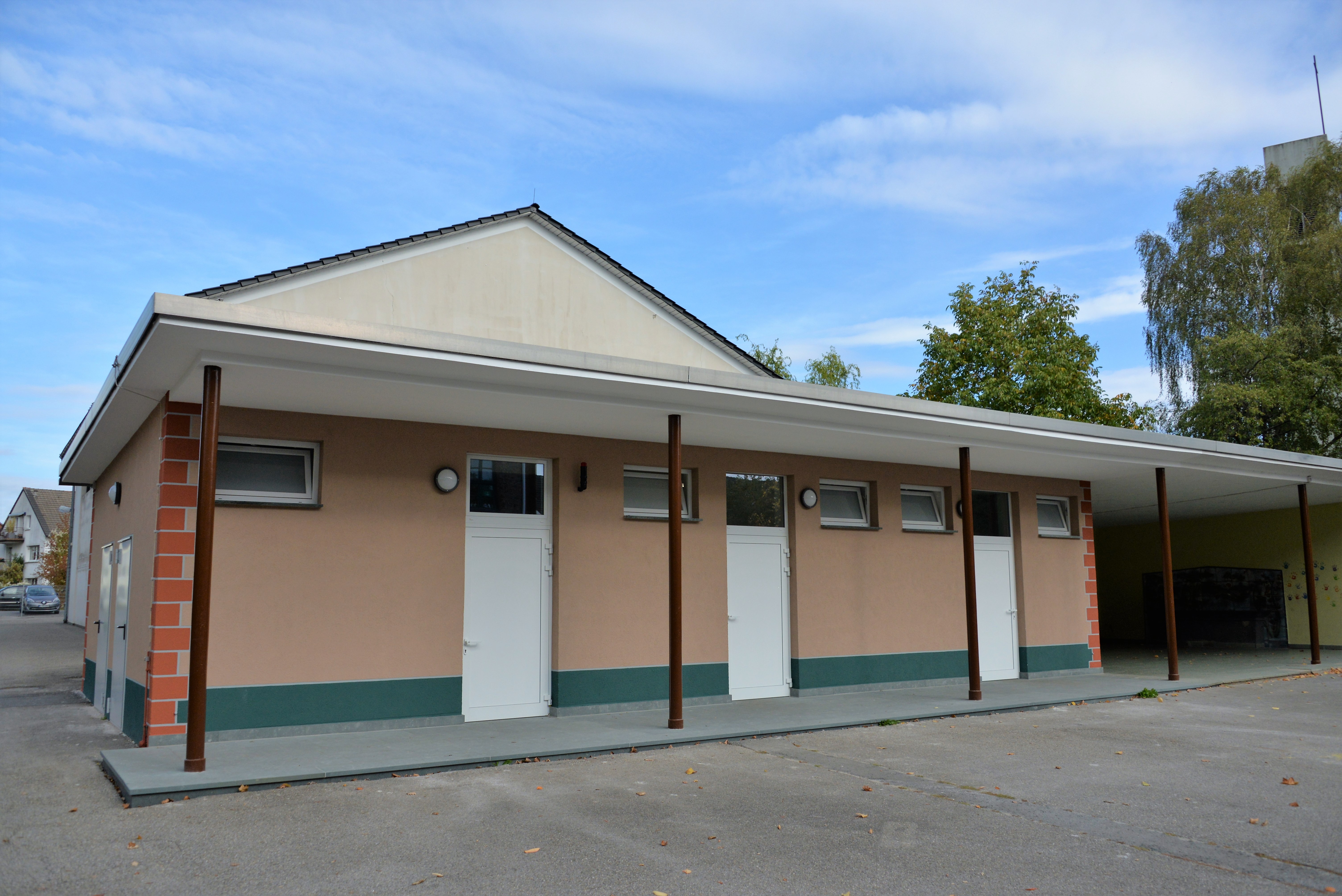 Foto des Toilettengebäudes der Grundschule am Hellweg am Standort Horster Berg nach erfolgter Renovierung. Die Außenansicht zeigt das Gebäude mit drei separaten überdachten Eingängen zu den Toilettenanlagen und eine neugestaltete Fassade in den Farben Altrosa und im Sockelbereich Petrol