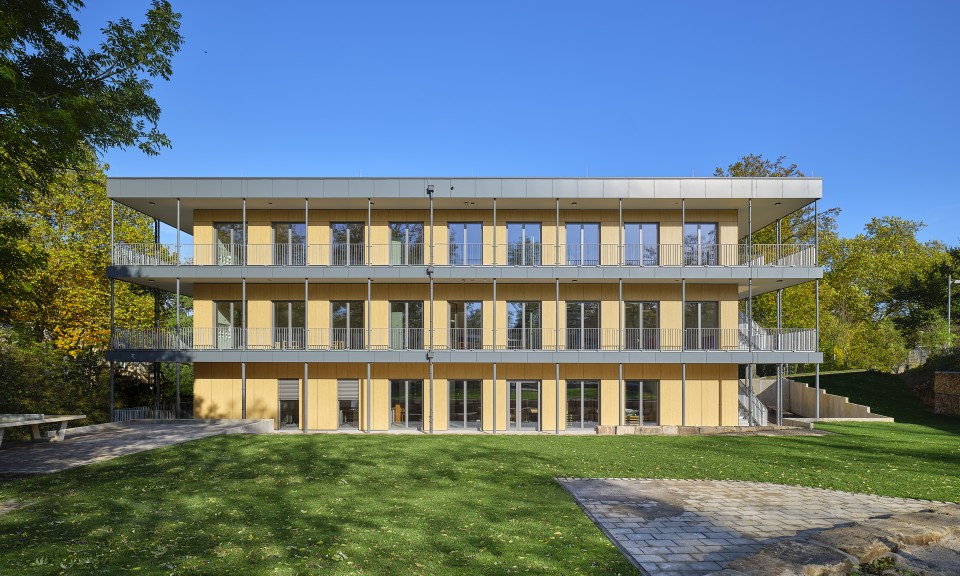 Das Foto zeigt die Fassadenansicht des dreigeschossigen Schulneubaus in Modulbauweise am Schulstandort Rosastraße 80 von Süden. Bodentiefe zweiflügelige Fenster und Türen verteilen sich gleichmäßig auf der Fassade, es sind zwei umlaufende Balkone zu sehen. Das Gebäude hat ein Flachdach und an den Kopfseiten Fluchttreppen. 