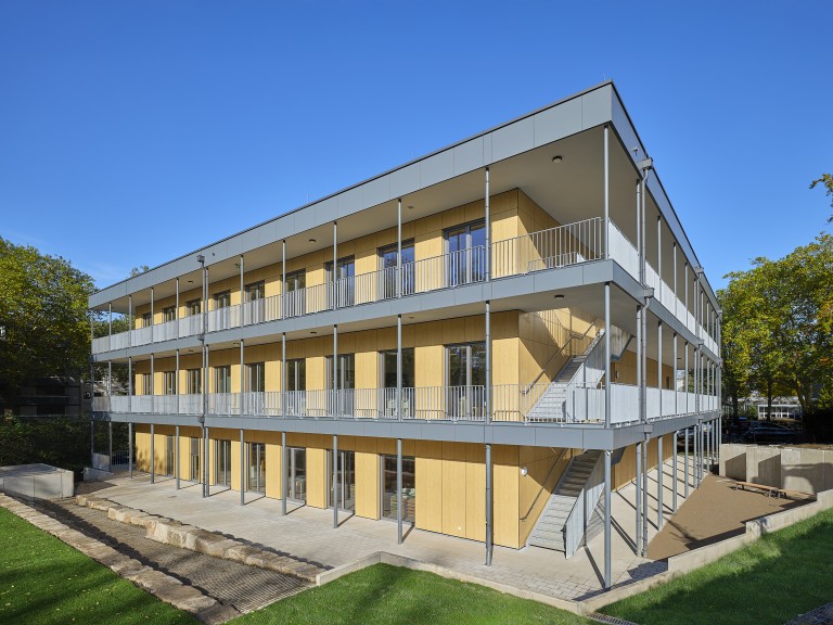 Das Foto zeigt die Fassadenecke des dreigeschossigen Schulneubaus in Modulbauweise am Schulstandort Rosastraße 80 von Südost. Bodentiefe zweiflügelige Fenster und Türen verteilen sich gleichmäßig auf der Fassade, es sind zwei umlaufende Balkone zu sehen. Das Gebäude hat ein Flachdach, an der rechten Kopfseite ist eine von zwei Fluchttreppen zu sehen. 