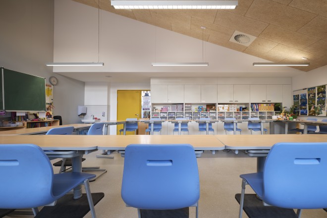 Auf dem Foto ist ein typischer und wiederkehrender Klassenraum im Erweiterungsneubau der Andreasgrundschule an der Von-Einem-Straße 56 in Essen-Rüttenscheid zu sehen. Die höhenverstellbaren Schultische -Stühle sind zu einem großen Viereck mit Ausrichtung auf eine Wandtafel zusammengestellt. In der Bildmitte ist ein weißer Einbauschrank, der mit offenen und geschlossenen Elementen versehen ist, zu sehen. Hier werden Lernutensilien untergebracht. Eine Besonderheit des Raumes ist die sehr hohe und schräge, der Dachform folgenden reversiblen Akustik-Decke. 