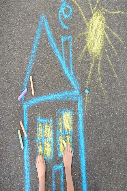 Foto: ein Kind zeichnet mit Kreide ein Haus auf dem Asphalt.