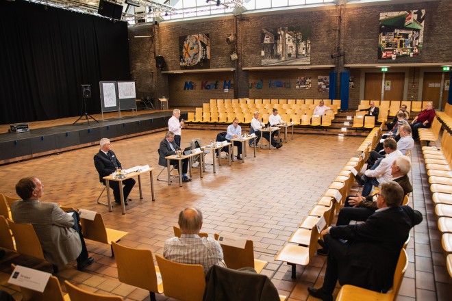 Foto: In der Mitte eines großen Saals sind sechspersonen an jeweils einem Tisch nebeneinander aufgereiht. Eine der Personen steht mit einem Mikrofon in der Hand. Mehrere Menschen sitzen um sie herum.