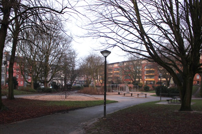 Leerer Spielplatz mit blattlosen Bäumen und einer bunten Häuserreihe im Hintergrund